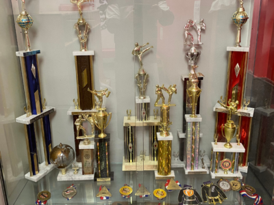 A display case at Zhang Sah martial arts school in Philadelphia showcasing karate trophies and medals earned across decades of competition, including World Tang Soo Do Championship titles.